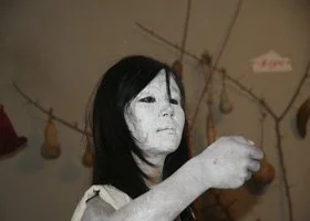 A person dressed as a ghost or zombie with white face paint, black hair, and dark clothing, holding a small object in their hand.