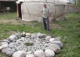 Man standing outside near a yurt, with a rock circle and small stones in the foreground.