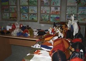 Table displaying various handmade crafts and knitted items, with drawings and more crafts on the wall in the background.