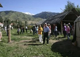 Group of people standing in a grassy field near traditional houses with mountains in the background.
