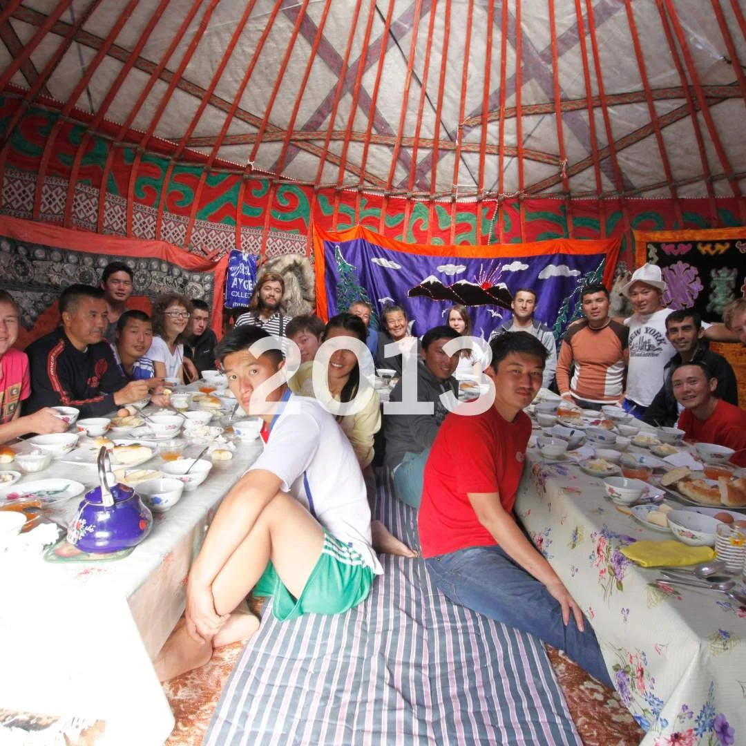 Group of people sitting and standing around a long table inside a decorated tent for a celebration, with dishes of food and drinks on the table. The photo has a large '2013' overlay.