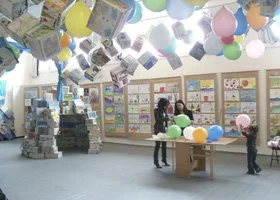 Children in a classroom decorated with balloons and hanging photos, with shelves filled with books and toys.