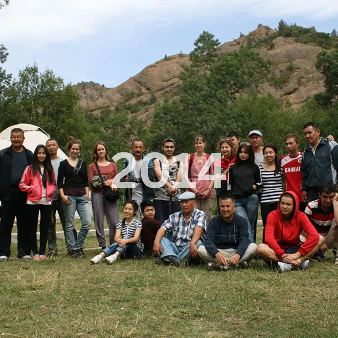 A group of about 16 people, including men, women, and children, are gathered outdoors on a grassy area with trees and rocky hills behind them. Some individuals are standing, while others are sitting on the ground. The photograph appears to be from the year 2014, according to the white text overlay.