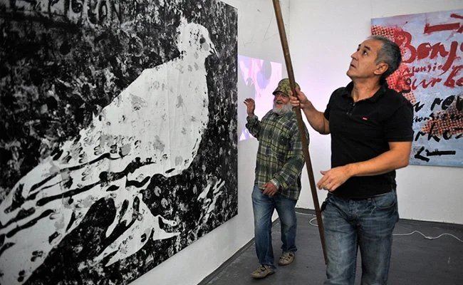 Two men observe a black and white artwork of a bird on a gallery wall, one holding a long stick or pointer.