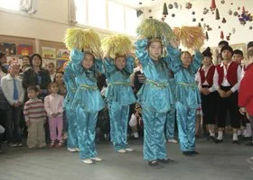 Three people dressed in blue traditional costumes with yellow headdresses performing a dance or parade indoors, with a crowd of spectators watching.