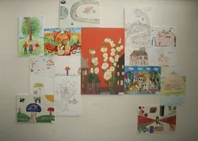 Collection of children's drawings and paintings displayed on a wall, featuring various colorful scenes and objects.