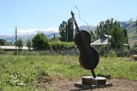 A sculpture of a person playing a cello made of metal with a wire for the bow, set outdoors on a grassy area with mountains and trees in the background.