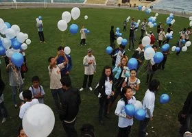 Group of people gathering outdoors, holding blue and white balloons, on a grassy area near a body of water.
