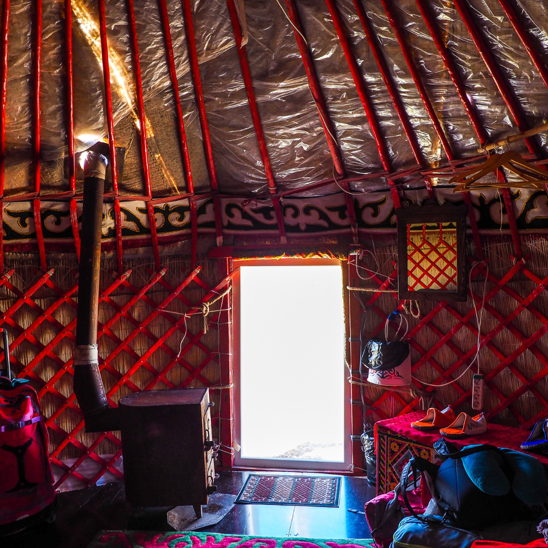 Interior view of a traditional yurt with a door letting in bright sunlight. The yurt has red wooden lattice walls and a stove pipe on the left. There are bags, shoes, and various items inside.
