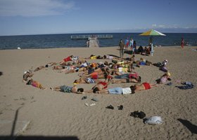 A group of people forming a large heart shape on a sandy beach near the ocean, with a pier and colorful umbrellas in the background.