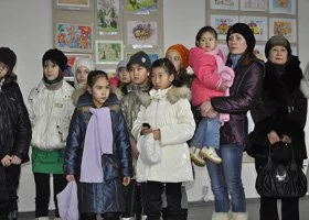 Group of children and adults standing together indoors, some holding bags or jackets, against a wall with colorful artwork.