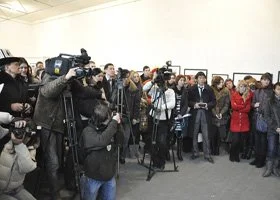 Group of journalists and photographers with cameras and microphones gathered at an indoor press event.