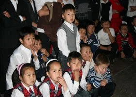 Group of young children sitting and standing together, some with surprised or curious expressions, in a crowded indoor setting.