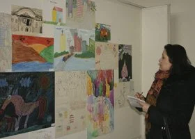 A woman standing in front of a wall displaying various colorful children's drawings and paintings.