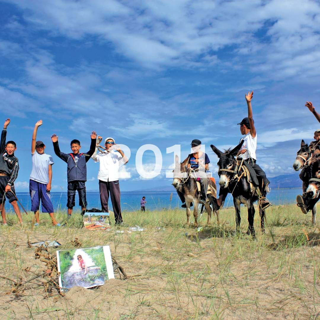 Children riding donkeys on a grassy beach, waving and smiling, with photos on the ground and a blue sky with some clouds above.