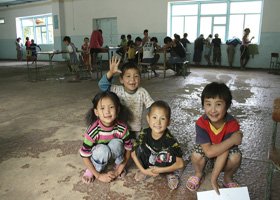 Four children sitting on the floor of a gymnasium, with a group of people in the background near large windows.