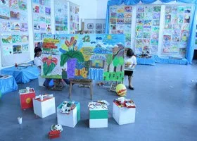 Children are viewing colorful painted panels and artwork displayed on tables and walls at an indoor art exhibit or classroom.