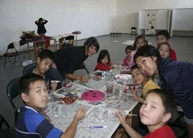 Children and adults are at a table making crafts or jewelry in a classroom.