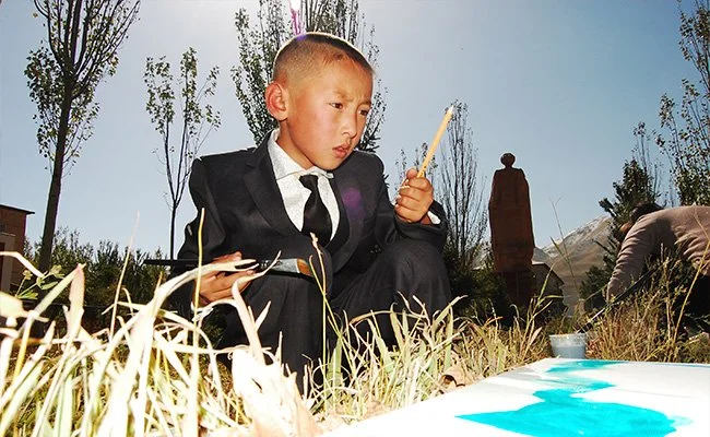 A young boy in a suit sitting outdoors on grass and holding a pencil, looking at a small pool of water.