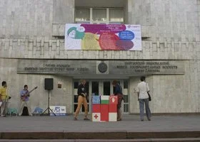 People gather around an information booth or display in front of a building with a sign, with a large colorful sign or banner above.