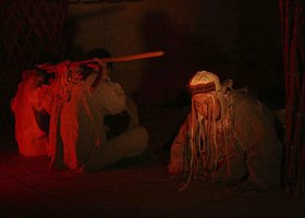 Two individuals sitting on the floor in a dimly lit environment with red lighting, one with a headband and long hair, the other wearing glasses, engaged in an activity.