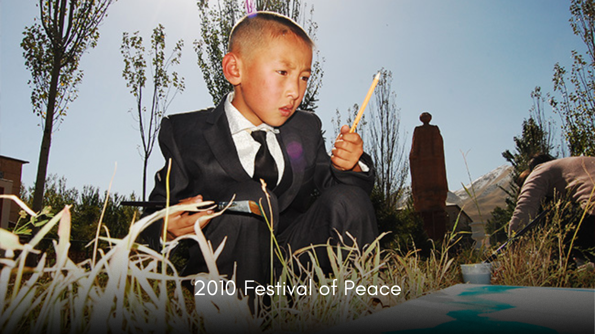A young boy in a suit and tie sits outdoors on the grass, holding a stick and looking at a paper or artwork. The background features trees, a chimney, and a mountain, with a person on the right side. The caption reads '2010 Festival of Peace'.