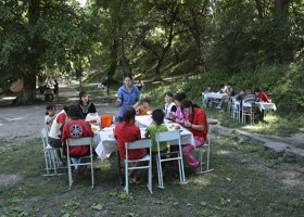Group of people dining outdoors at a round table in a wooded area.