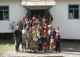 Group of children and adults standing in front of a white building with blue window frames, some children sitting, others standing, posing for a photo.