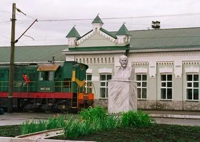 A green train near a historic building with white walls and green roofs, and a stone bust sculpture in front of the building.
