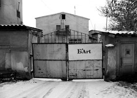 A black and white photo of a large metal gate with the word 'B'Art' on it, in a run-down area with old buildings and fences surrounding the gate.