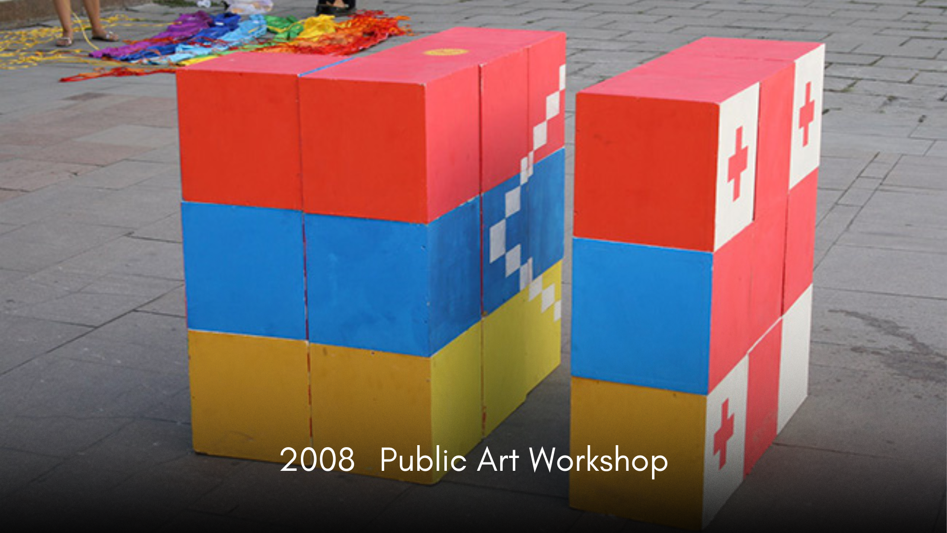 Colorful public art installation made of painted large wooden blocks arranged in a cube, with a palette of red, blue, yellow, and white, depicting pixelated designs including a heart and a cross, on a paved outdoor area during daytime.