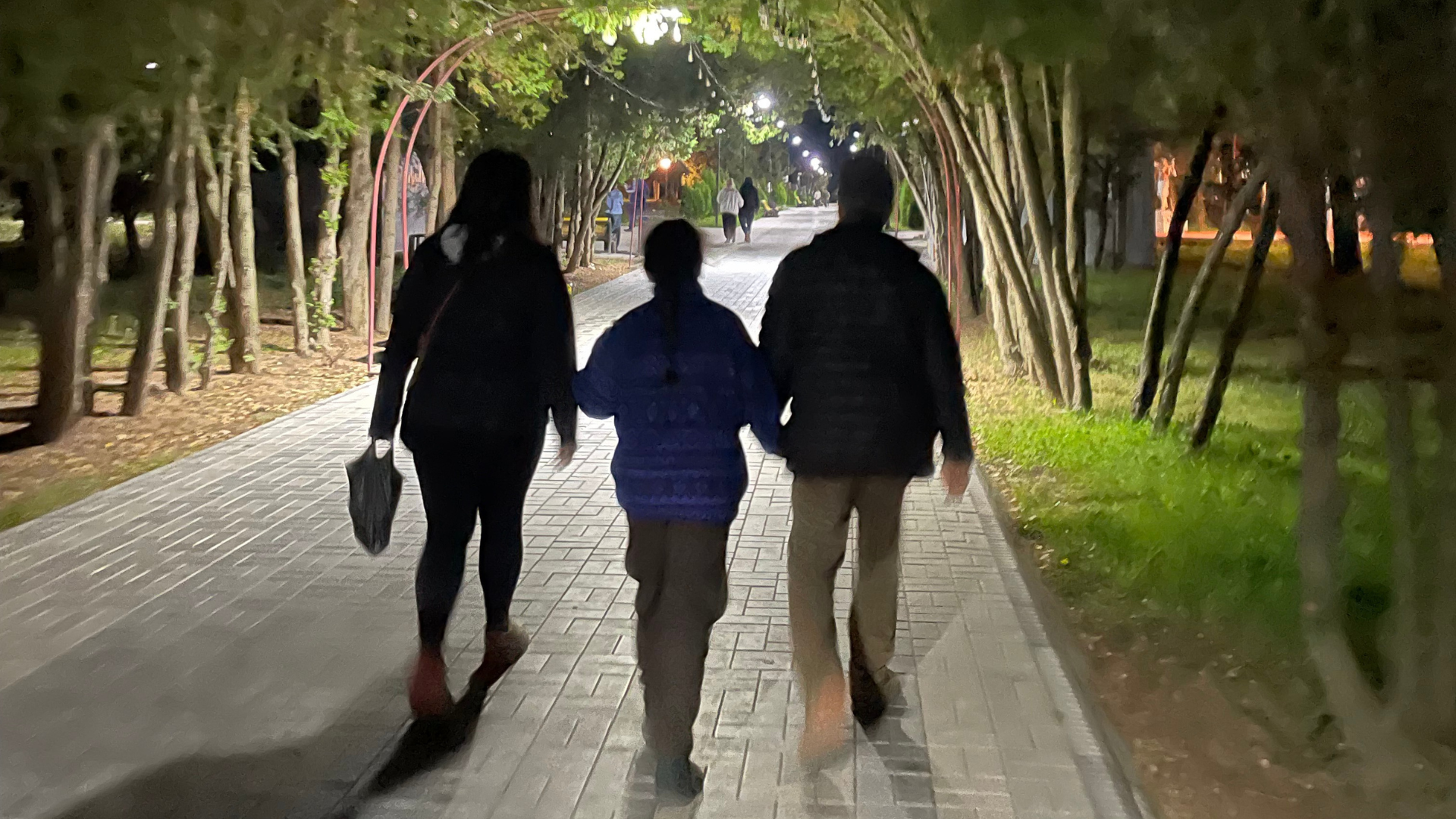 Three people walking hand in hand through a lit park tunnel at night, surrounded by trees and illuminated street lights.