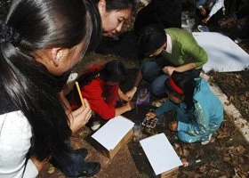Group of children and adults gathered closely outdoors, engaging in an activity with notebooks and a tablet on the ground.