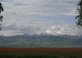 Landscape with snowy mountains in the background, green hills in the midground, and a field in the foreground, with cloudy sky overhead.