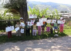 A group of children holding signs standing outdoors under a tree, with mountains and houses in the background.