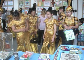 Group of young girls in gold dresses at an indoor event or exhibition, some holding shopping bags, with display tables and colorful decorations in the background.