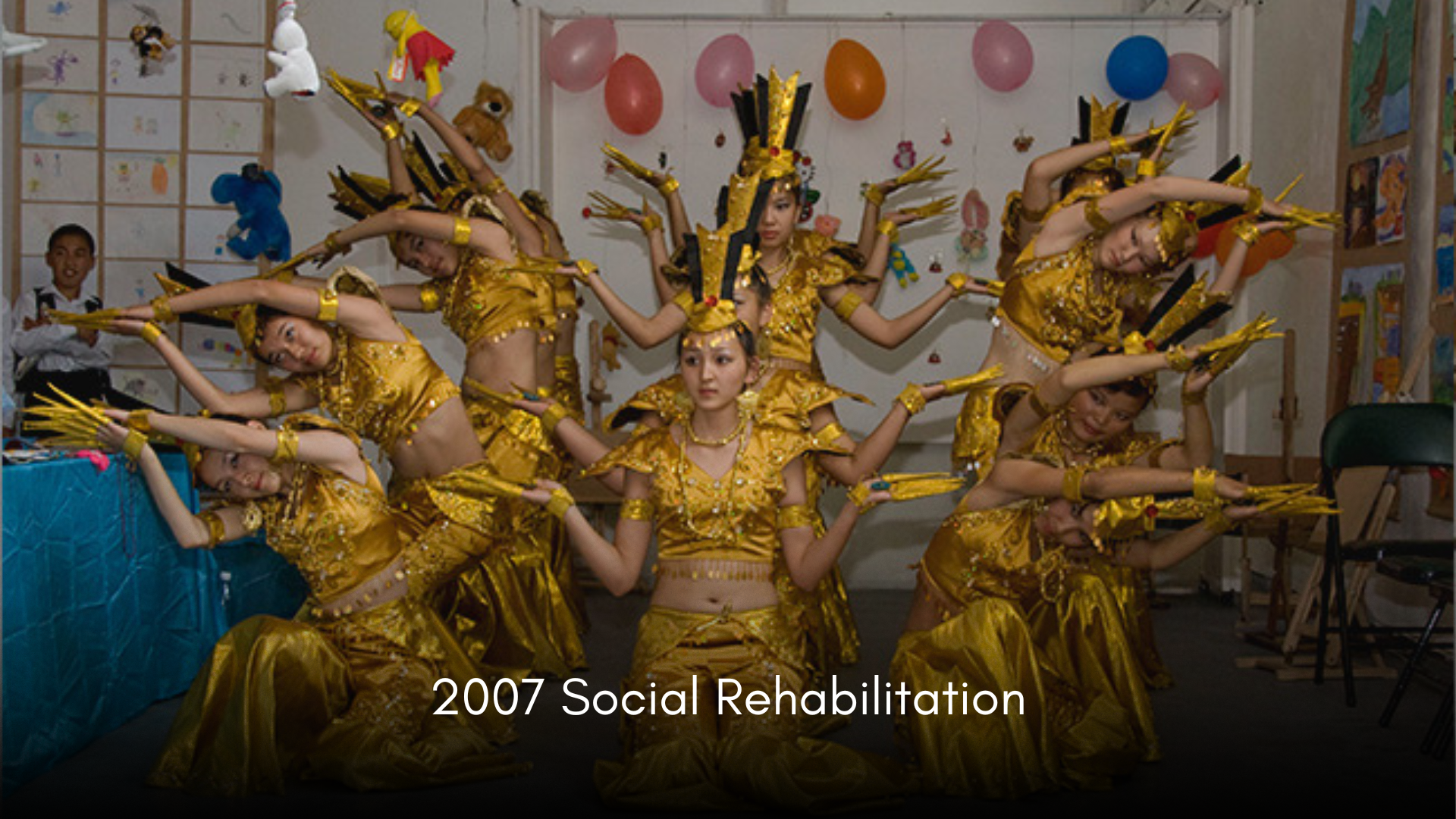 Group of young girls performing a traditional dance in golden costumes with headpieces and accessories, in an indoor setting decorated with balloons and drawings, with a few children watching in the background. Caption: 2007 Social Rehabilitation.