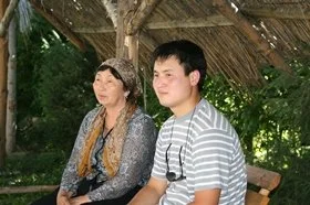 A woman and a young man sitting outdoors under a thatched roof, surrounded by green trees.
