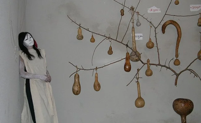 A mannequin dressed as a ghost with a white sheet and black hair, standing beside a wall with hanging branch-like structures decorated with various gourd-shaped objects and tags, possibly part of an art display or installation.
