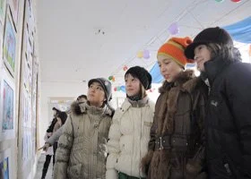Group of four women wearing winter coats and hats looking at artwork on a wall in an indoor exhibition.