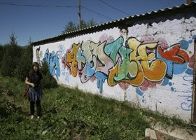 A girl standing next to a graffiti-covered wall outdoors on a sunny day.