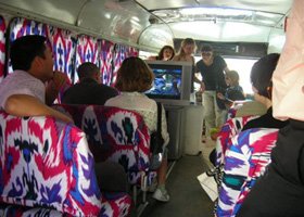 Inside a bus with passengers seated, colorful patterned seats, and a few people standing near the front over a monitor.