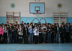 A large group of students standing together in a school gymnasium in front of sports equipment, including a basketball hoop, gymnastic rings, and wall-mounted climbing frames.