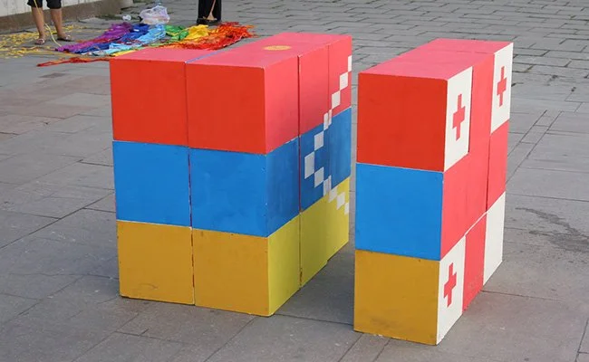 Two sets of stacked blocks painted to resemble the flags of China and Switzerland, placed on a sidewalk. The blocks have colorful pixel art designs.