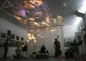 A group of people in an art gallery with projected clouds on the ceiling and walls, some seated and some standing, with artwork displayed on the walls.
