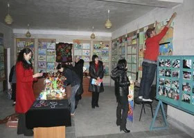 People in a classroom observing a woman pointing at a display board, with others at a table of crafts. The room has colorful decorations and photos on the walls.