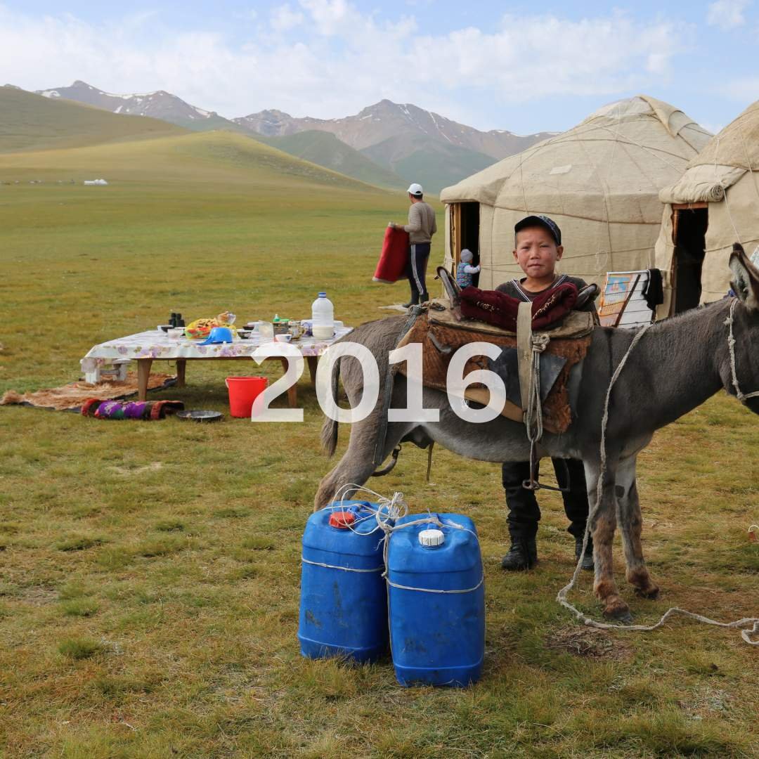 A young boy standing next to a donkey with blue water containers in a rural grassy field. Two yurts and a table with food and supplies are in the background, with mountains in the distance. The image has the year "2016" overlaid.