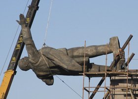 A large sculpture of a man lying down, being lifted or positioned by a crane, with scaffolding nearby against a blue sky.