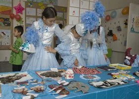 Three children in light blue costumes with white and blue feathered headpieces looking at a table with cookies and crafts during a festive event.