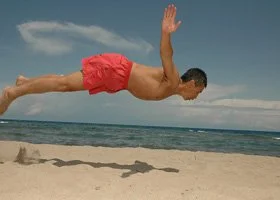 A man performing a dive into the water at the beach, with his body extended horizontally and arms outstretched behind him